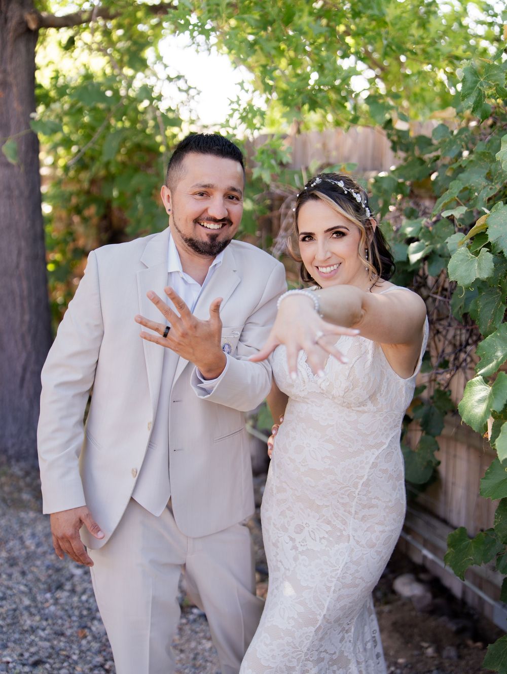 A wedding couple in light colored attire showing their rings outdoors with greenery and trees in the background.