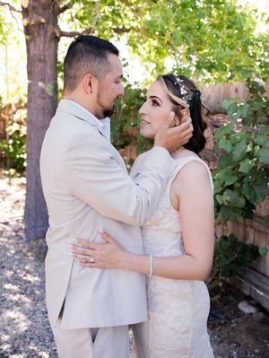 A romantic outdoor wedding moment between a couple wearing a light tan suit and white lace dress beneath trees in a garden.