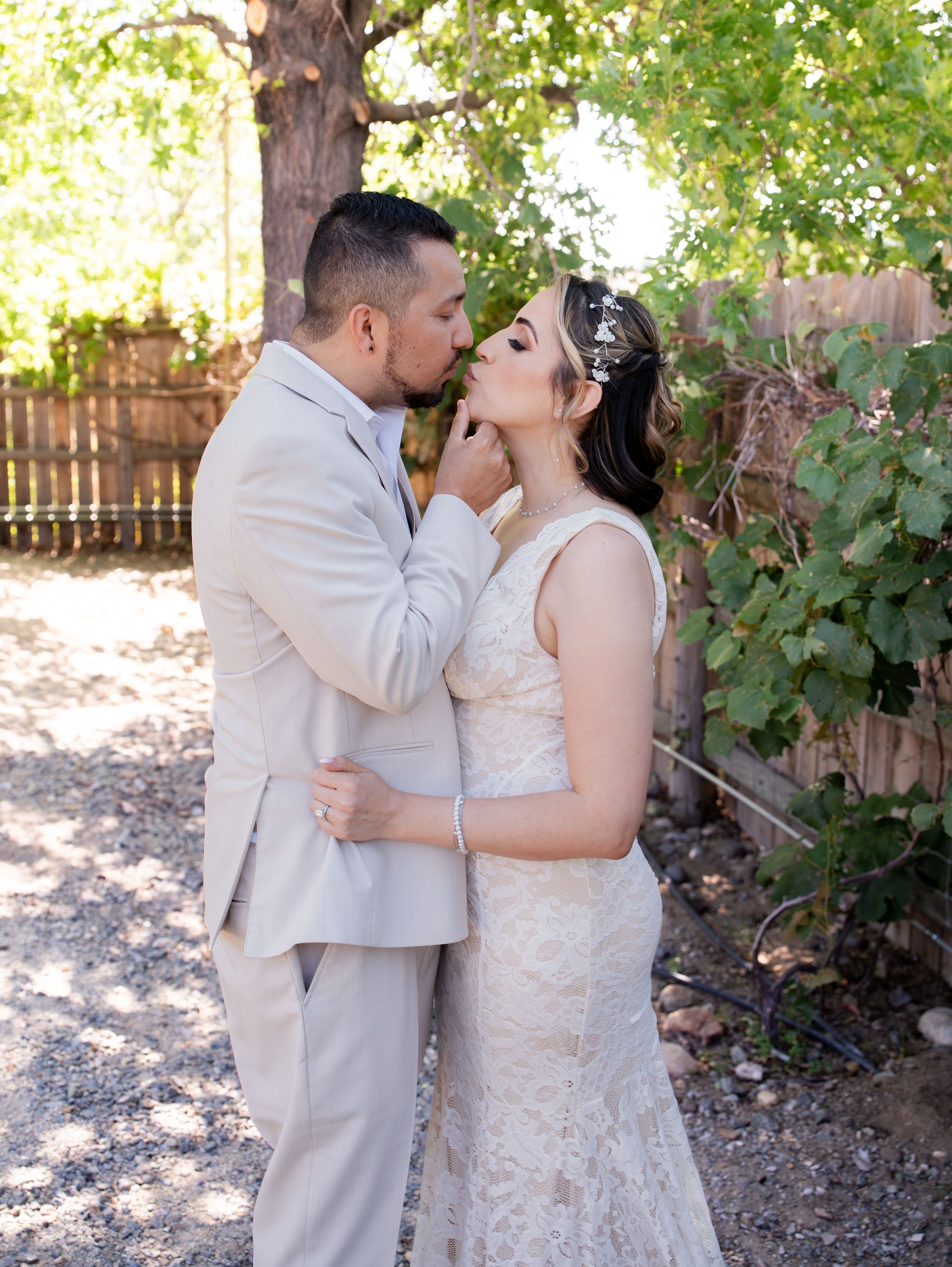 A couple in wedding attire share a romantic kiss under a lush green grape arbor in an outdoor garden setting.