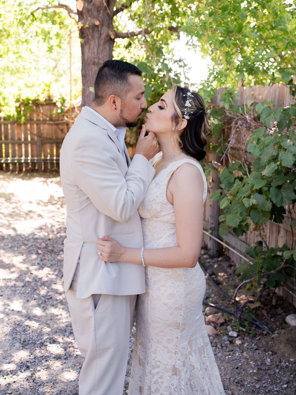 A couple in wedding attire share a romantic kiss under a lush green grape arbor in an outdoor garden setting.