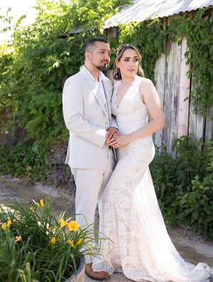 Bride in lace dress and groom in cream suit embrace in garden setting with yellow daylilies and wooden fence.