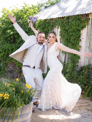 A joyful couple strikes playful poses in their wedding attire outside a rustic barn with climbing vines and yellow flowers.
