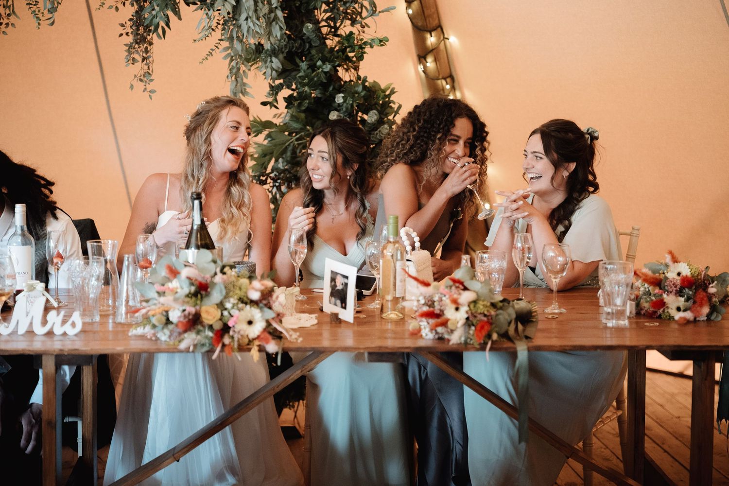 Wedding party shares laughter at rustic reception table with turquoise dresses under string lights and greenery.