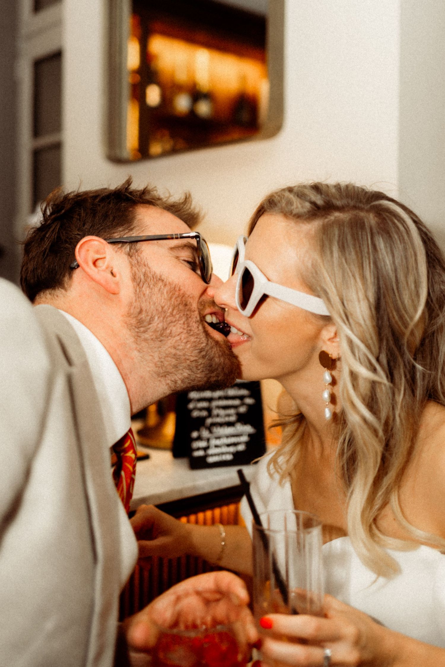 A bride in white dress and sunglasses kisses groom in gray suit at wedding reception while holding drinks.