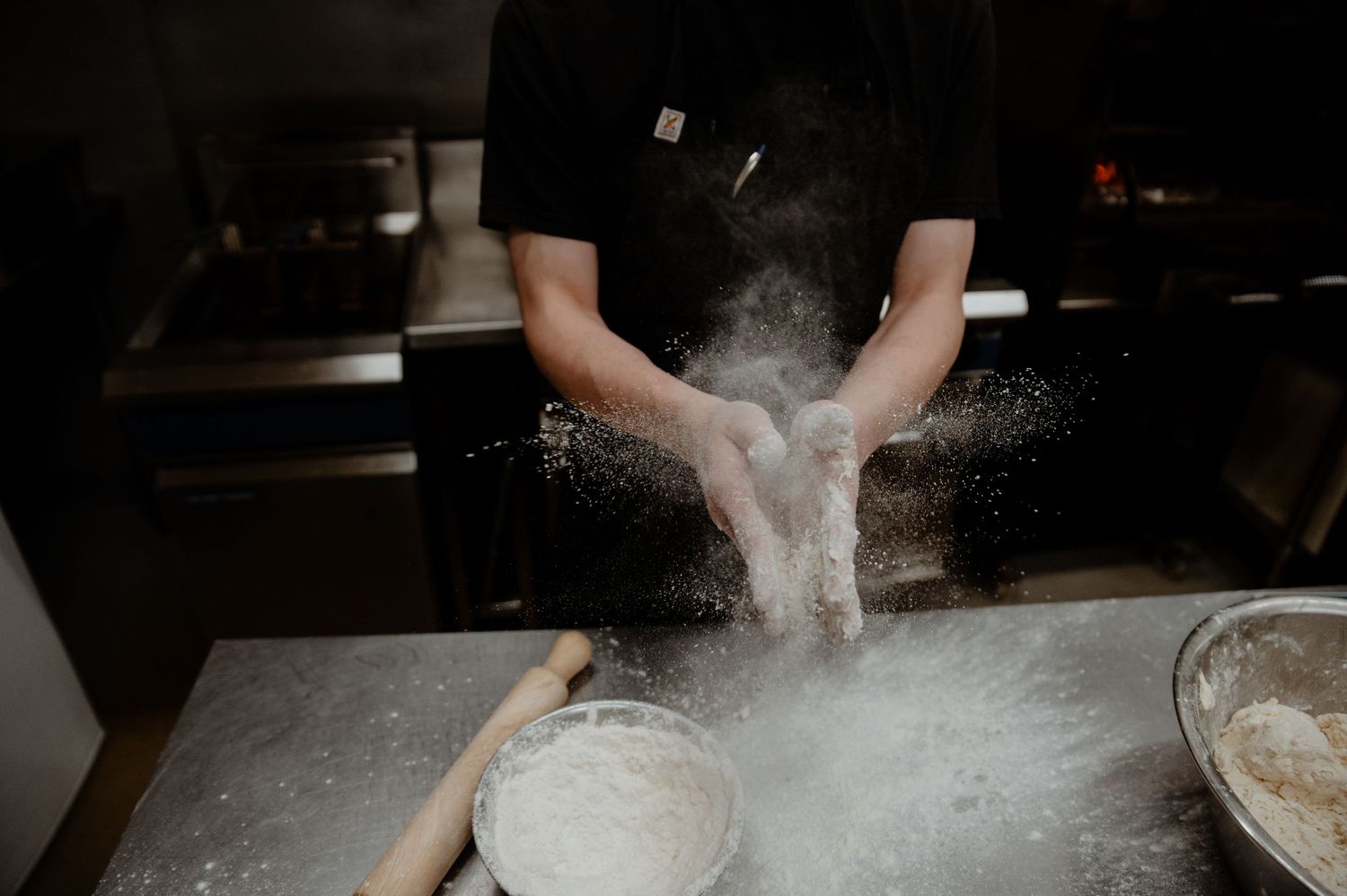 Chef's hands clapping flour into the air while kneading dough on a floured work surface in a professional kitchen.