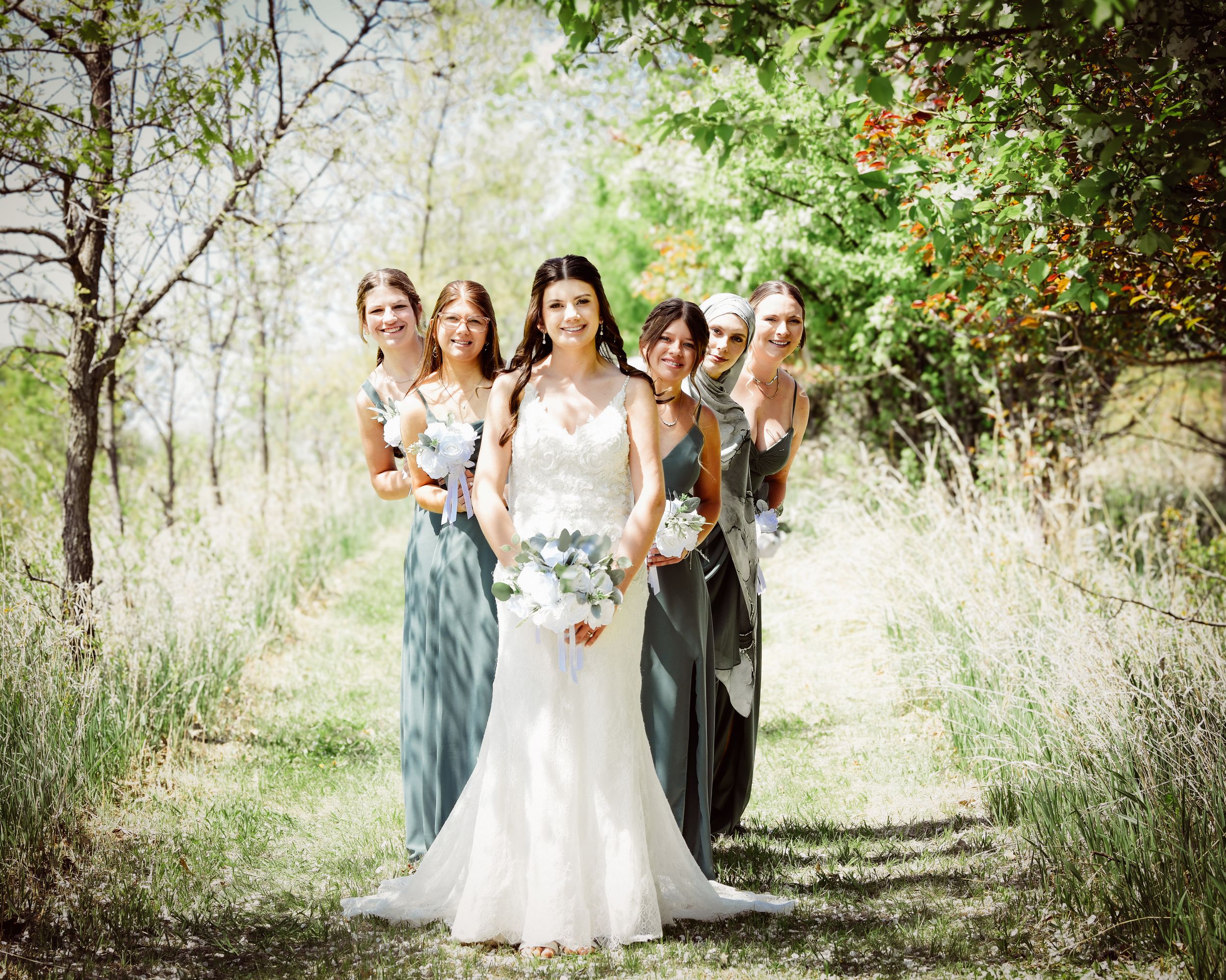 Wedding party stands among lush green trees in elegant attire on a sunny afternoon.