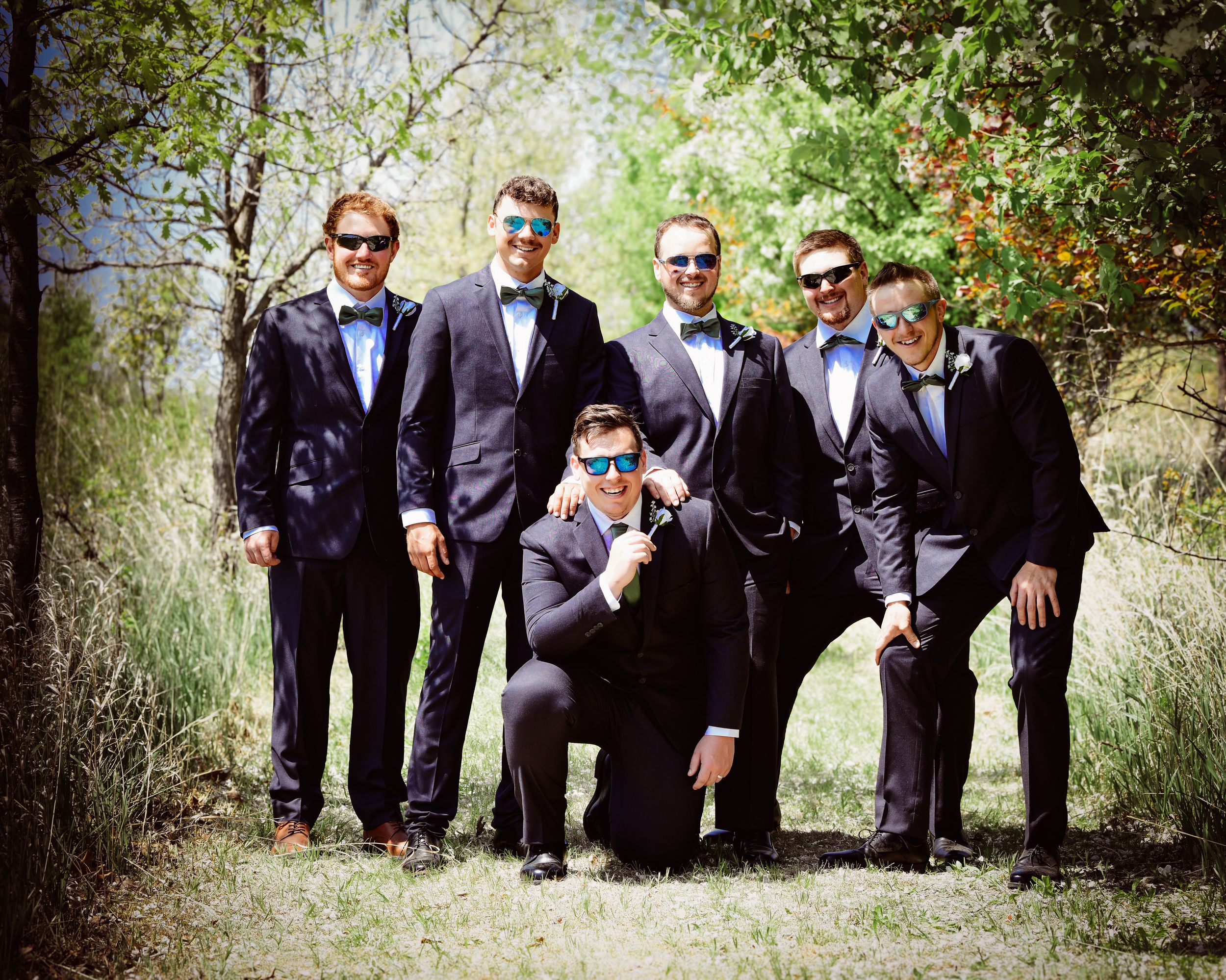 Groomsmen in dark suits and sunglasses pose casually in an outdoor garden setting.