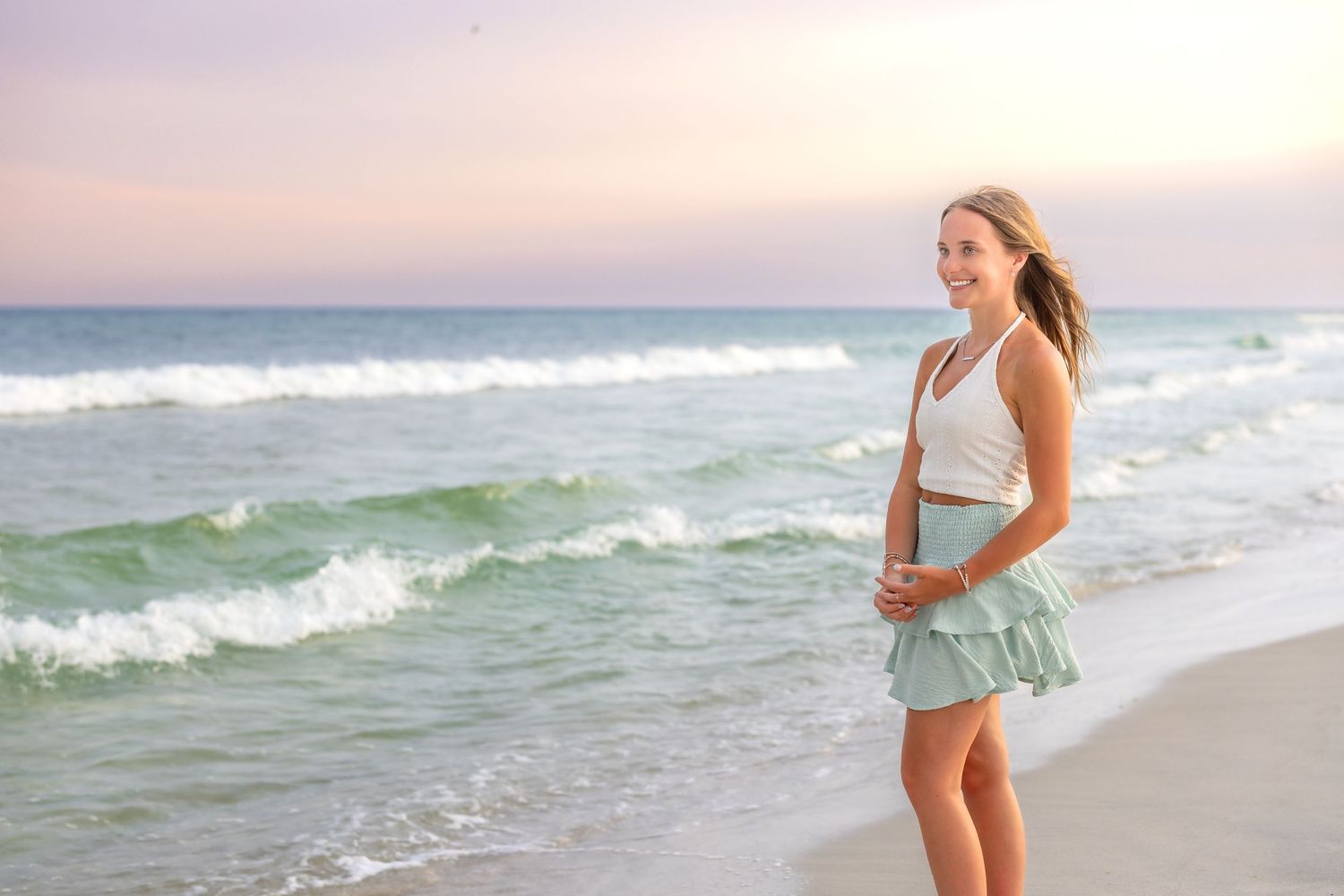 A person in flowing dress walks along a scenic beach at sunset as waves crash against the shore.
