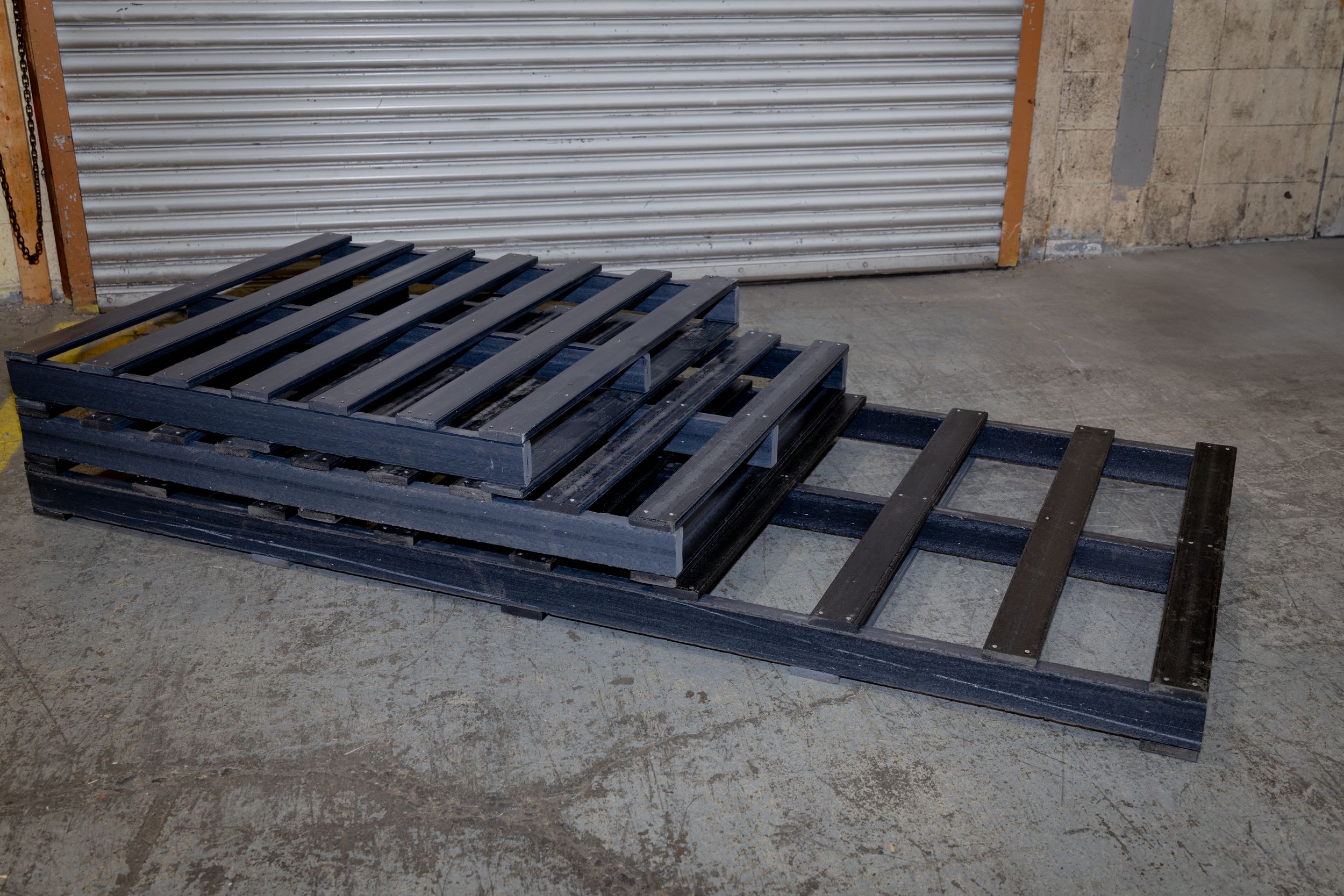 Five black wooden pallets positioned against a wall with corrugated metal doors in an industrial setting.