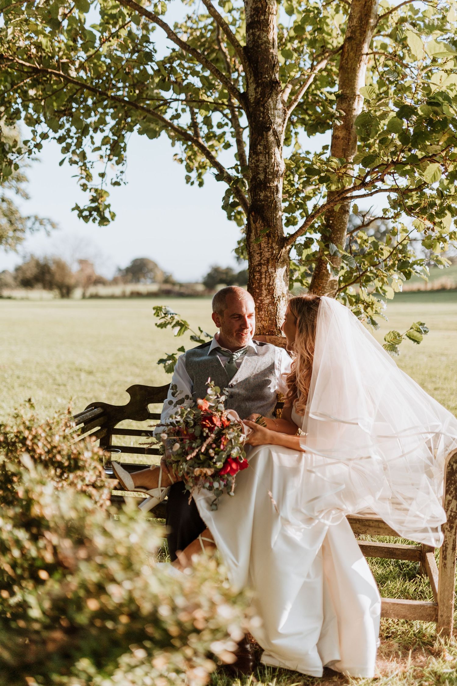 A romantic outdoor wedding photoshoot near a wooden fence and trees with a flowing white dress in warm sunlight.