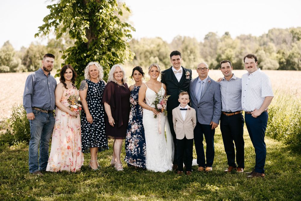 Wedding party and guests standing together in a field with tall grass and trees in the background on a sunny day.