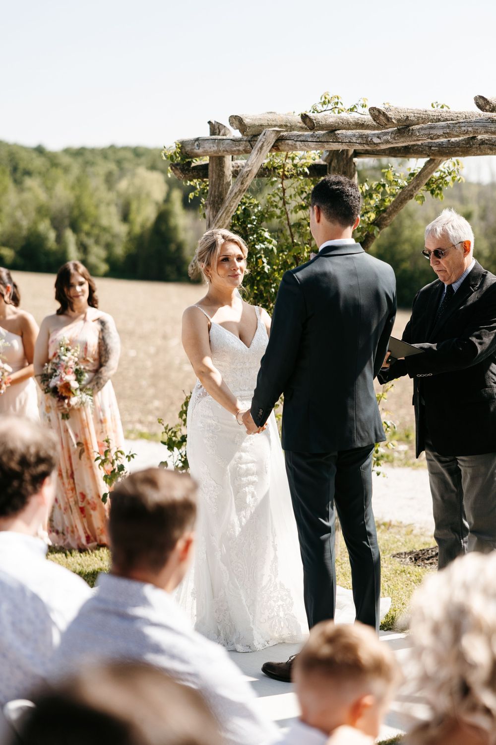 An outdoor wedding ceremony takes place under a rustic wooden arch in a field surrounded by trees and guests.