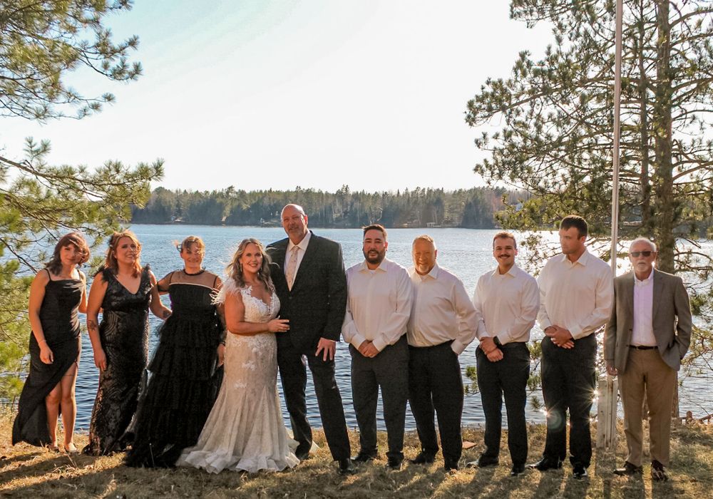 A wedding party poses together on a scenic lakeside cliff with pine trees and sparkling water in the background.
