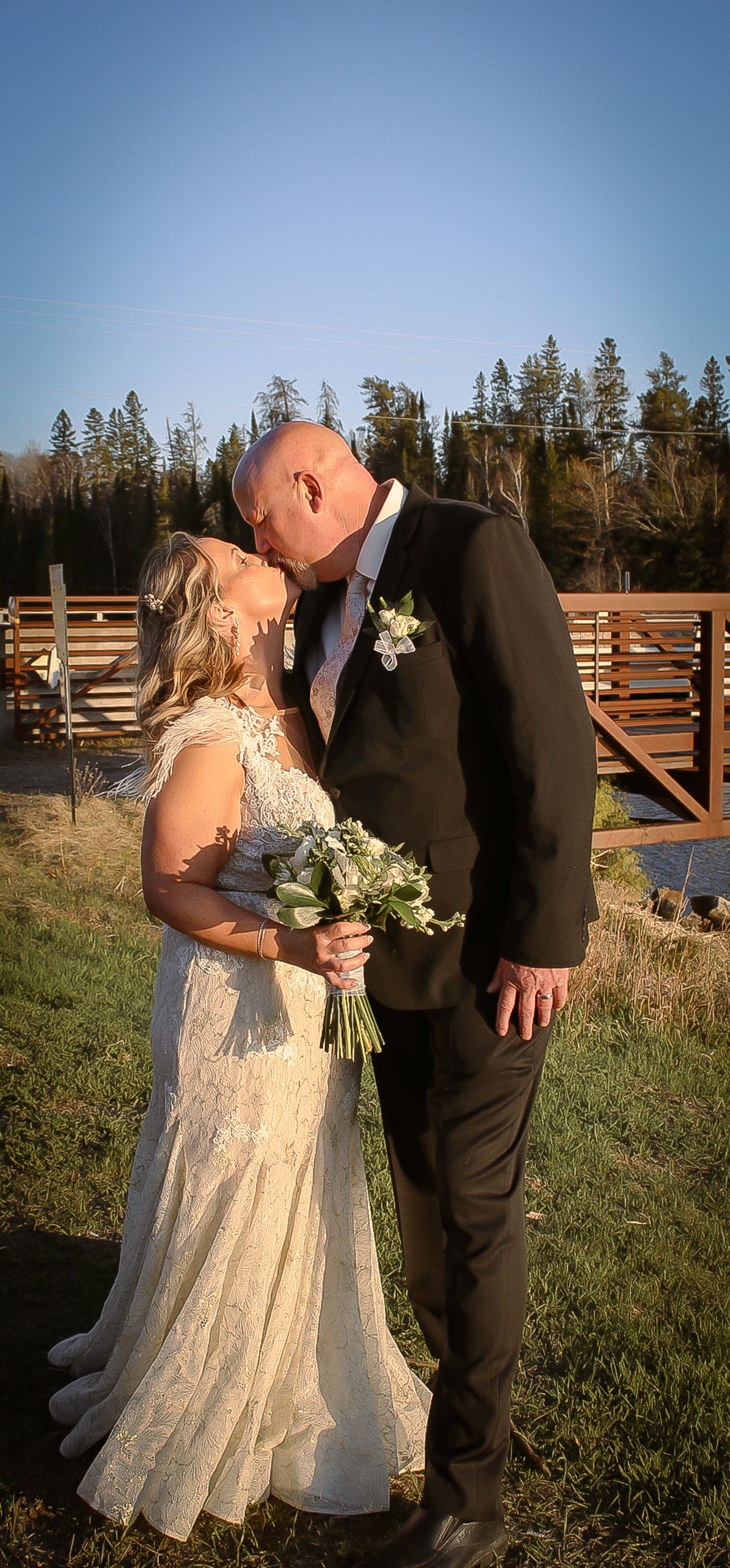 A romantic outdoor wedding couple embrace on a sunny day with wooden fencing and evergreen trees in the background.