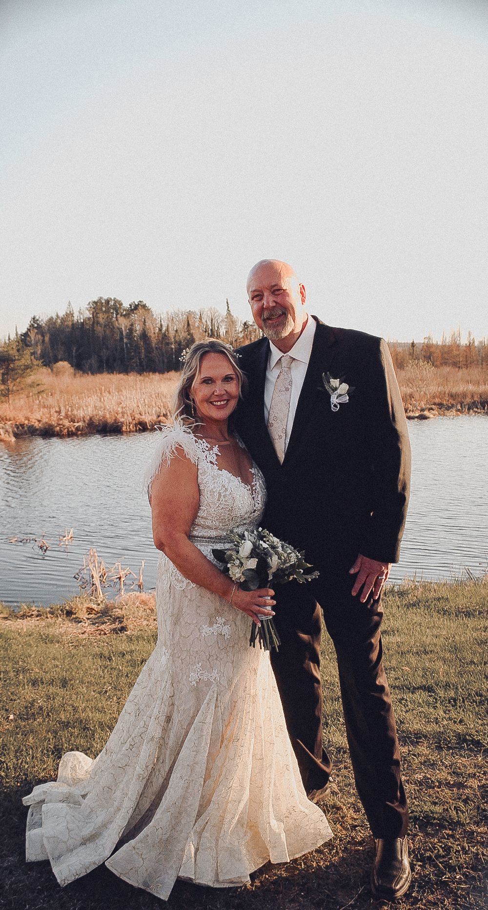 A couple poses together on their wedding day near a lake at sunset, with trees in the background.