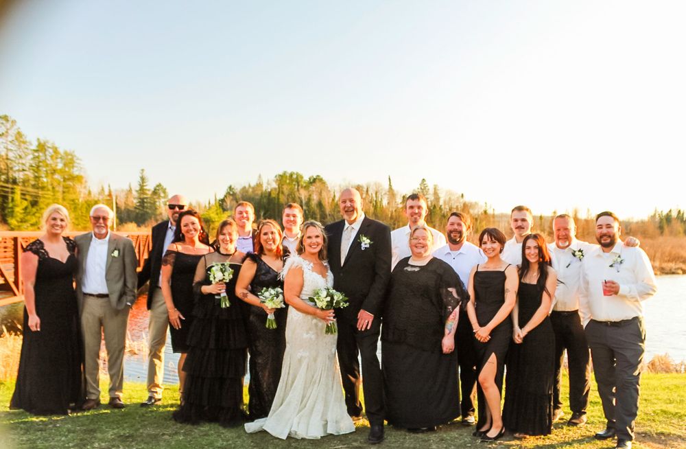 A wedding party in formal attire poses outdoors during golden hour on a grassy hill against a bright sky.