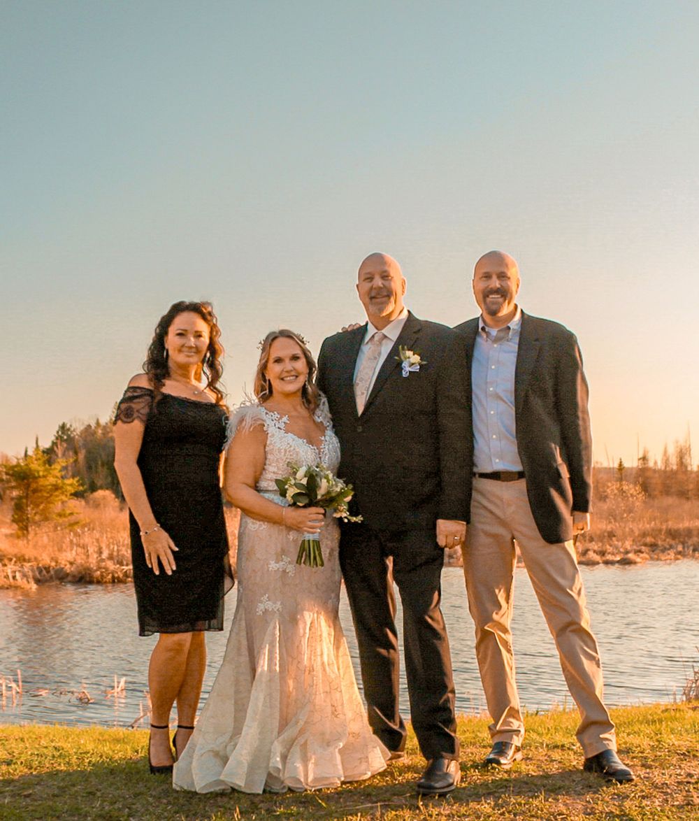 A group of people in formal attire pose together outdoors at sunset by a lake with fall foliage in the background.