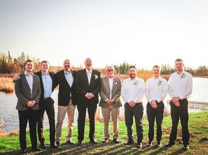 Wedding party members stand together on a field during sunset in black suits with the groomsmen wearing brown shoes.