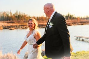 A couple in wedding attire walks along a lakeside beach during golden hour sunset.