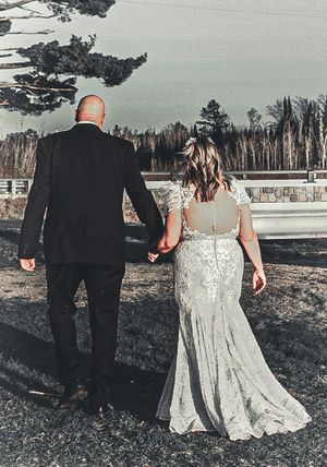 A couple in wedding attire hold hands while walking outside near a lake with trees in the background on an overcast day.