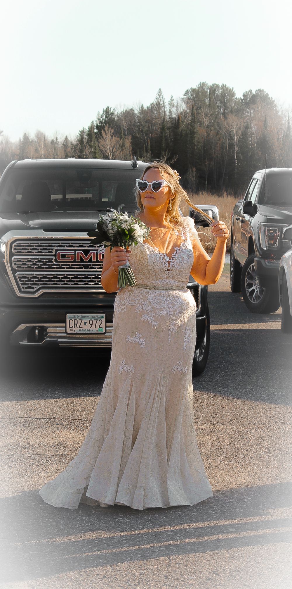 A wedding party member in a formal white lace dress poses beside a GMC truck in a sunlit outdoor setting.