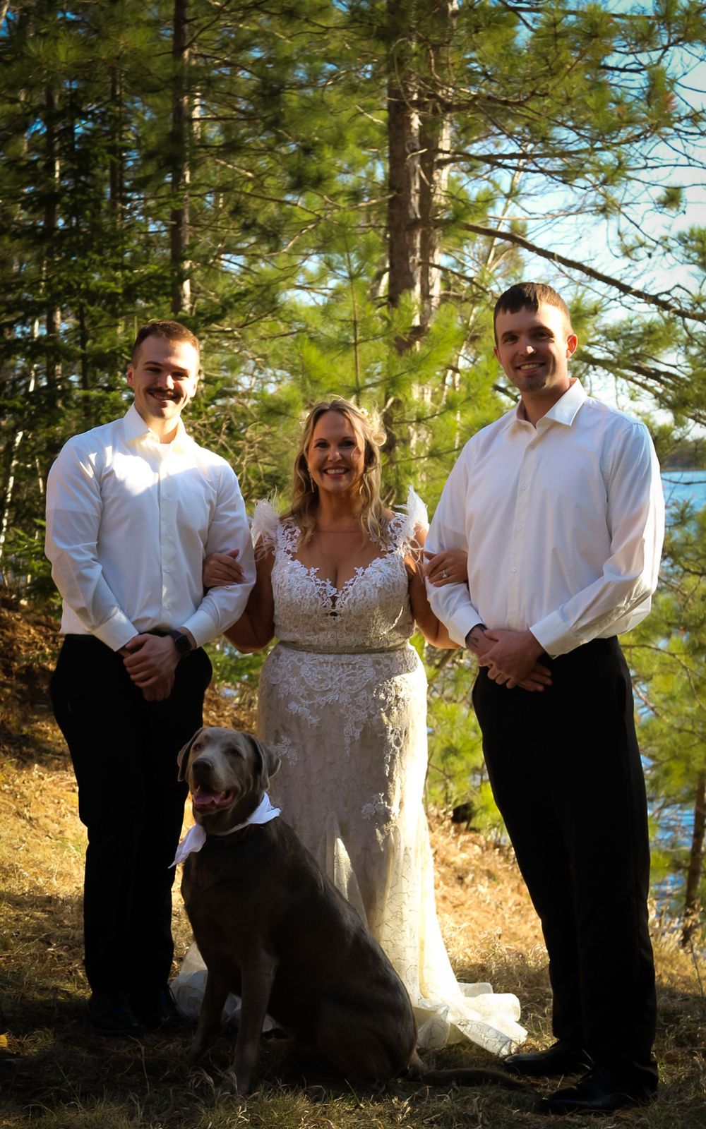 Three people in white attire pose outdoors with a black dog against a forested lakeside background.
