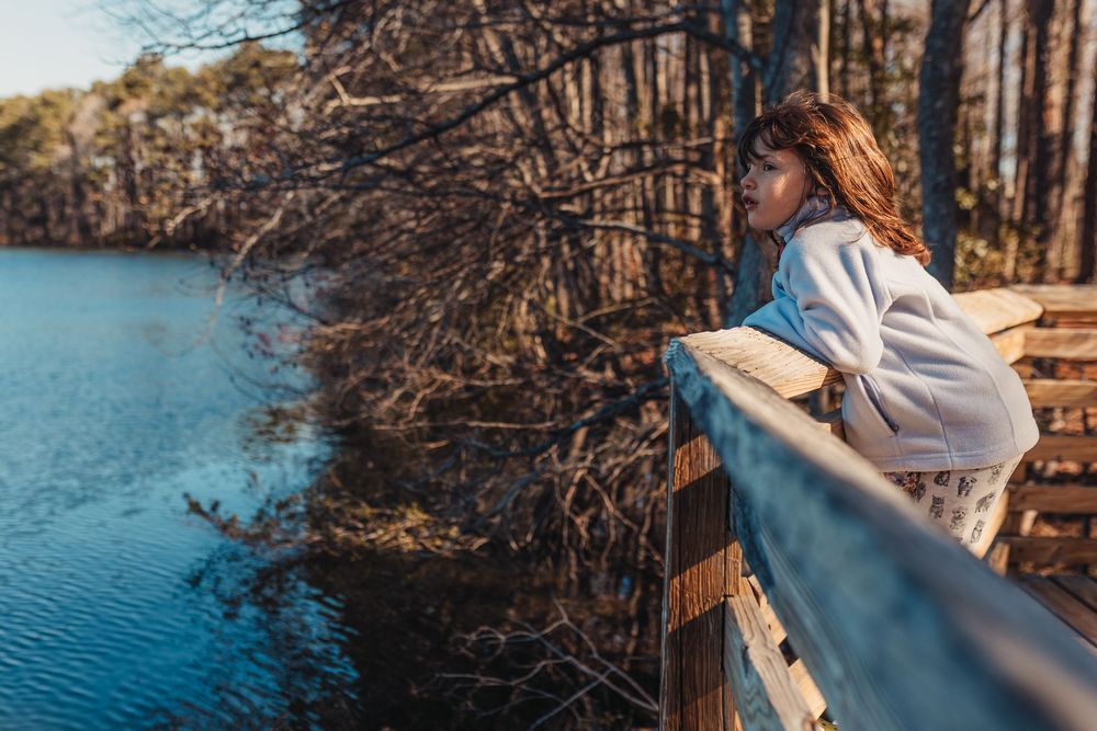 Little girl sitting on wooden dock overlooking calm lake waters surrounded by bare trees on a peaceful autumn day. Whitney Popp