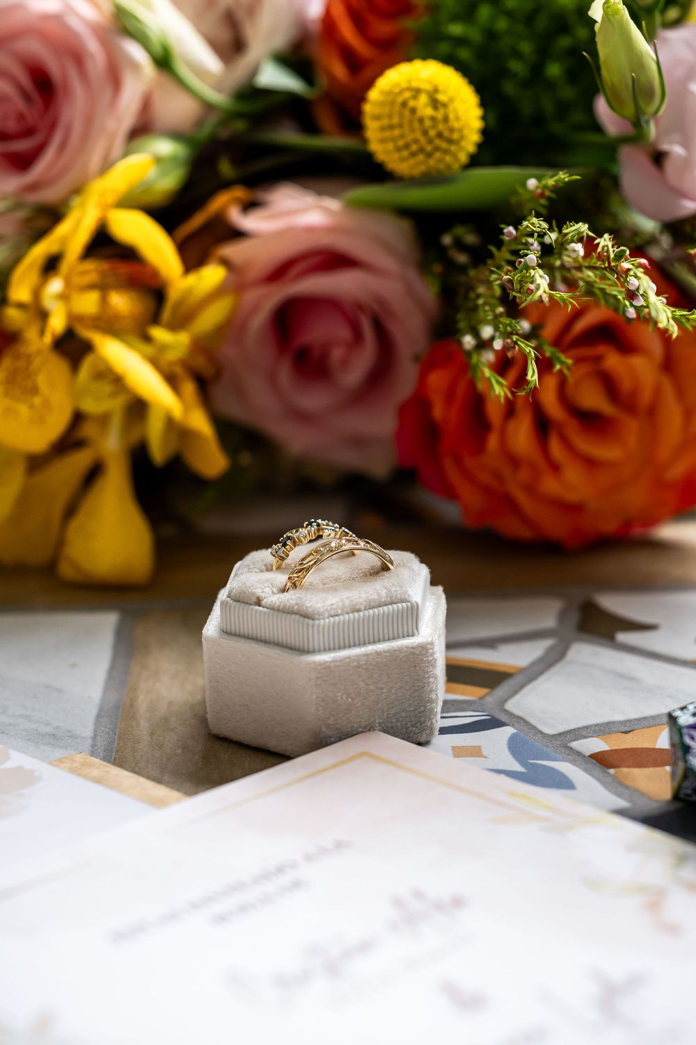 Wedding rings displayed in gray jewelry boxes among pink roses and yellow flowers on white surface.