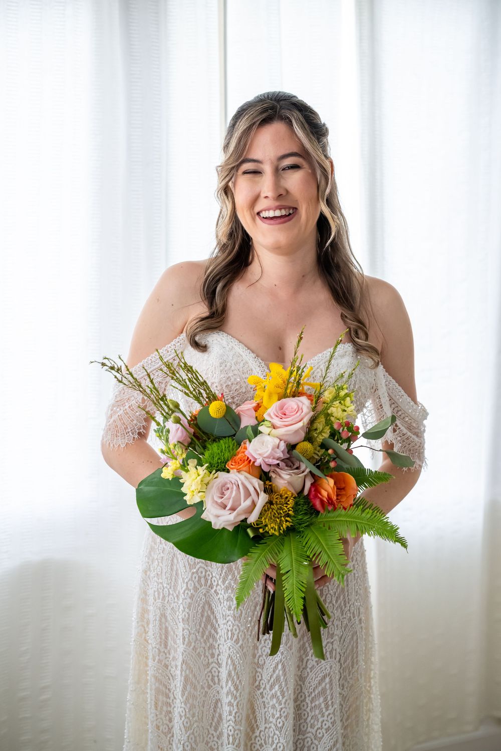 A joyful person in a white lace wedding dress holds a vibrant spring bouquet with roses, sunflowers and greenery.