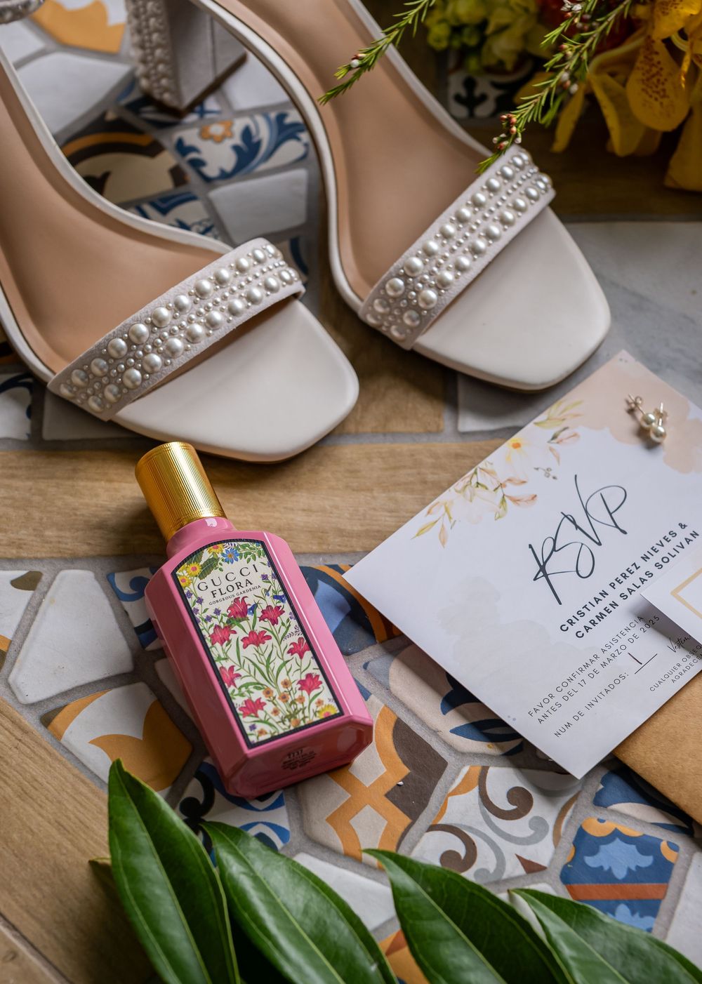 Wedding invitation cards and elegant bridal shoes with pearl embellishments arranged on a wooden surface with floral perfume.