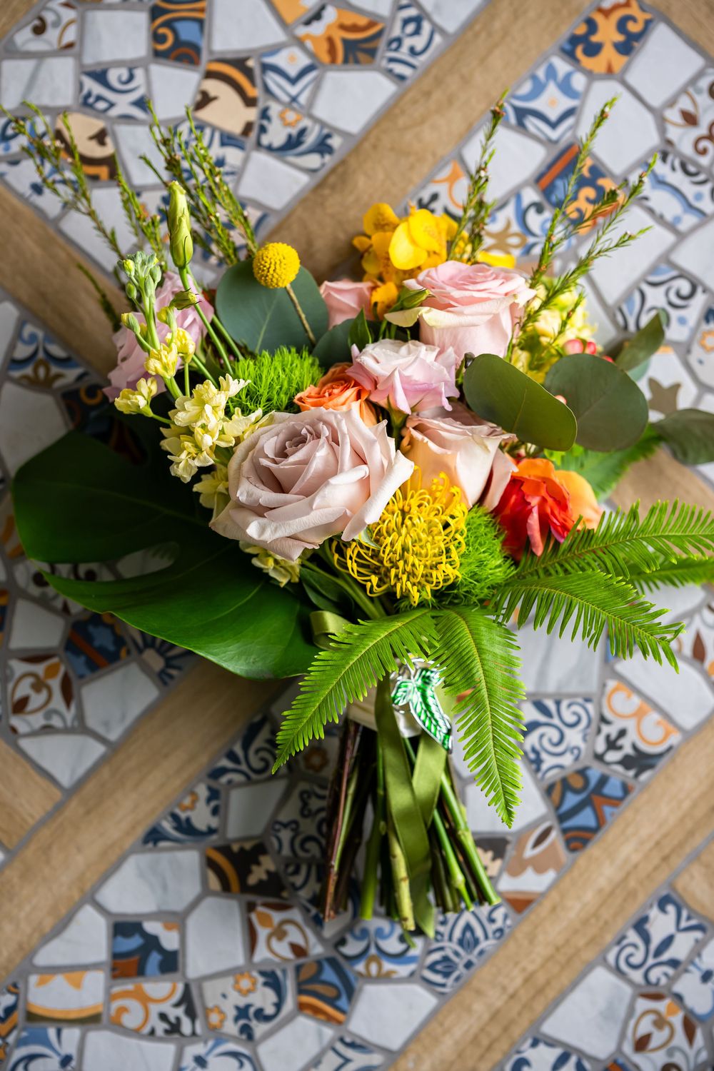A vibrant bridal bouquet with pink roses, yellow flowers and greenery arranged on decorative tiled surface.