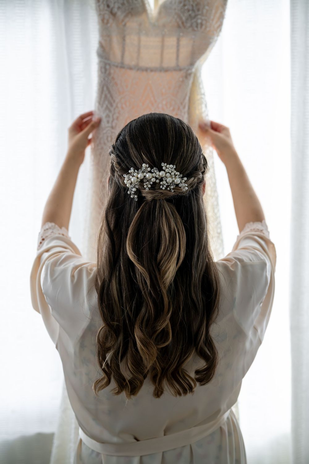 Back view of bride in white robe holding up wedding dress, showing elegant hairstyle with crystal hairpiece.
