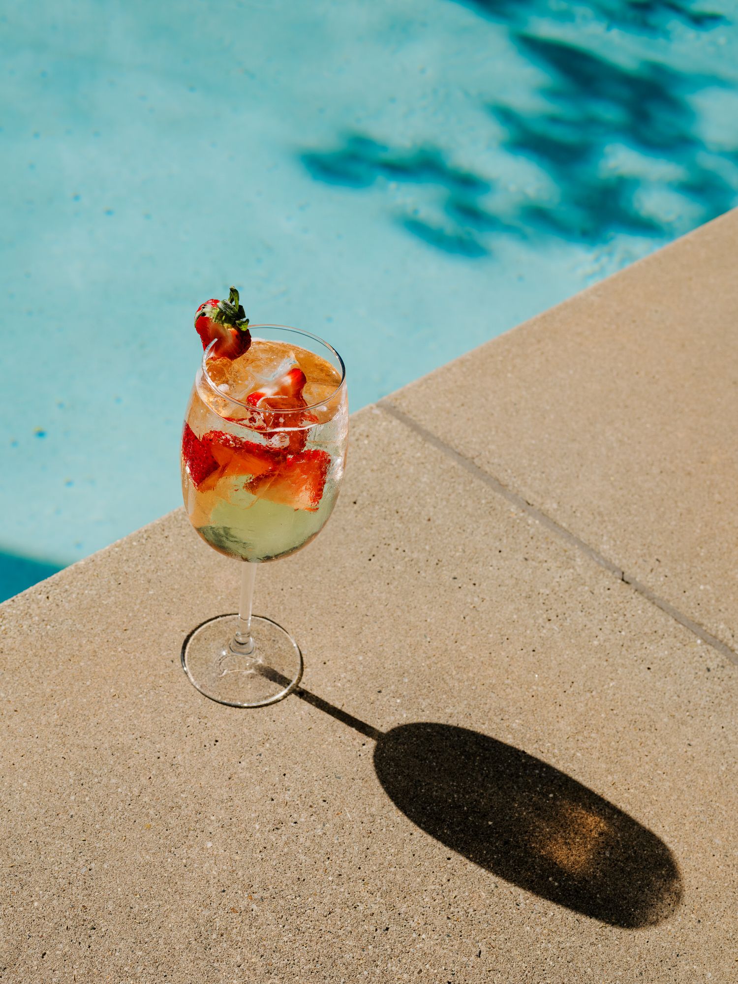 A colorful cocktail garnished with strawberries sits on a pool edge casting a long shadow in bright sunlight.