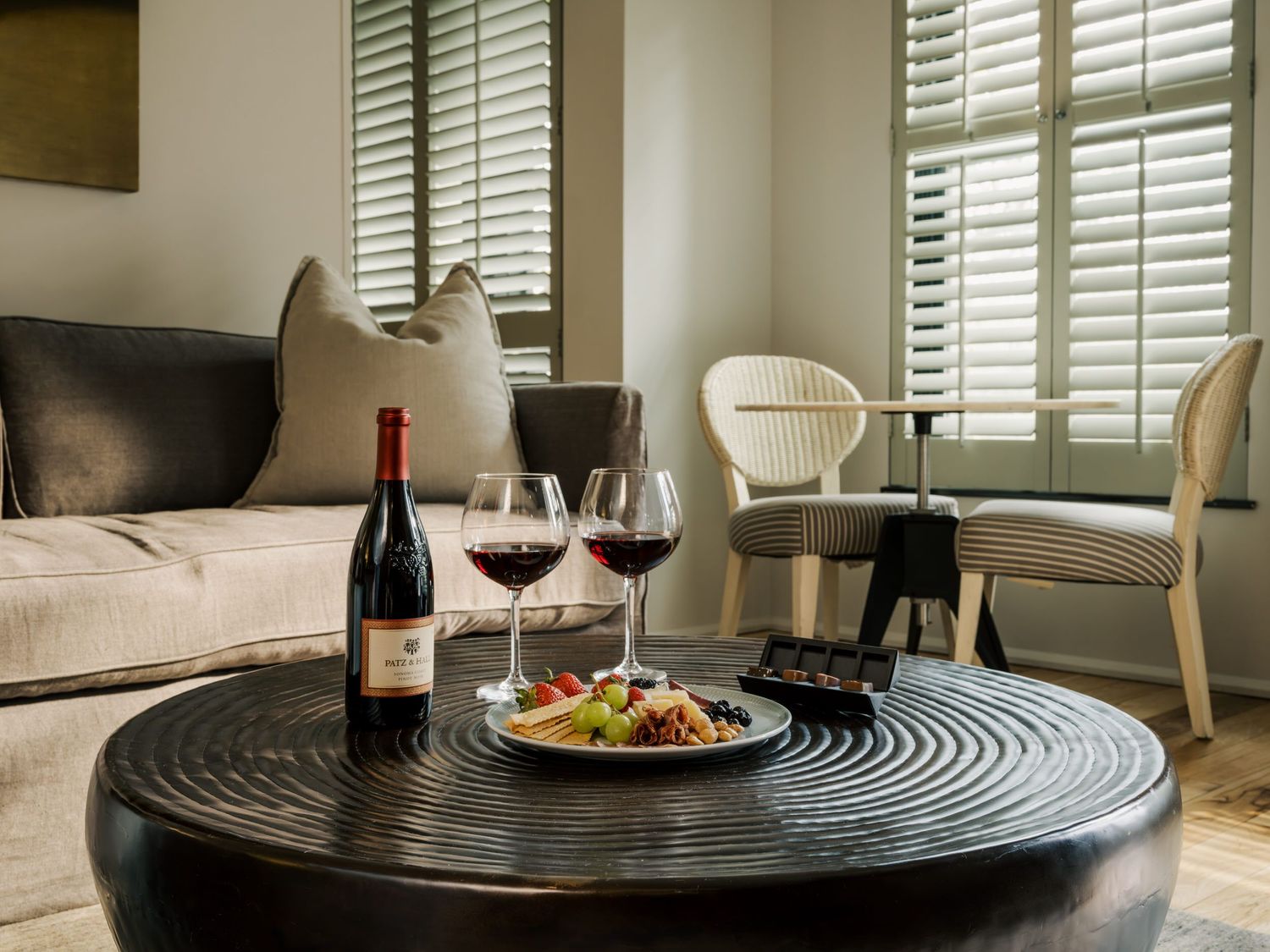 A modern dining setup with wine glasses and plates on a circular patterned placemat near window shutters.