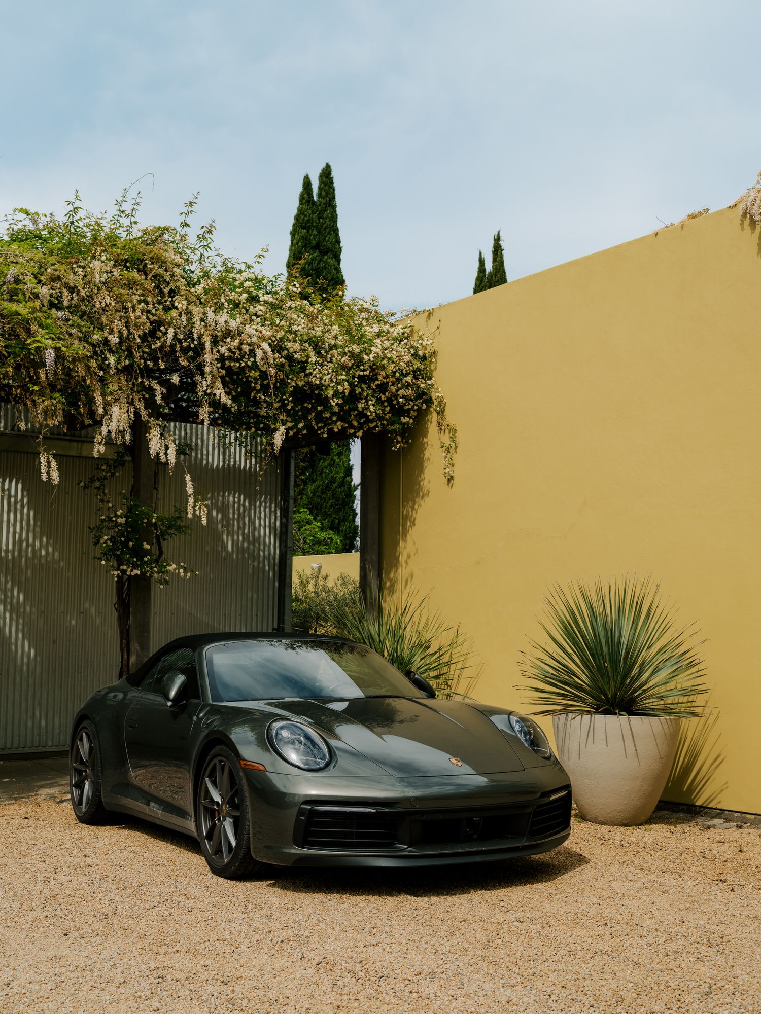 A sleek dark gray Porsche 911 parked in a Mediterranean-style courtyard with cypress trees and climbing vines.