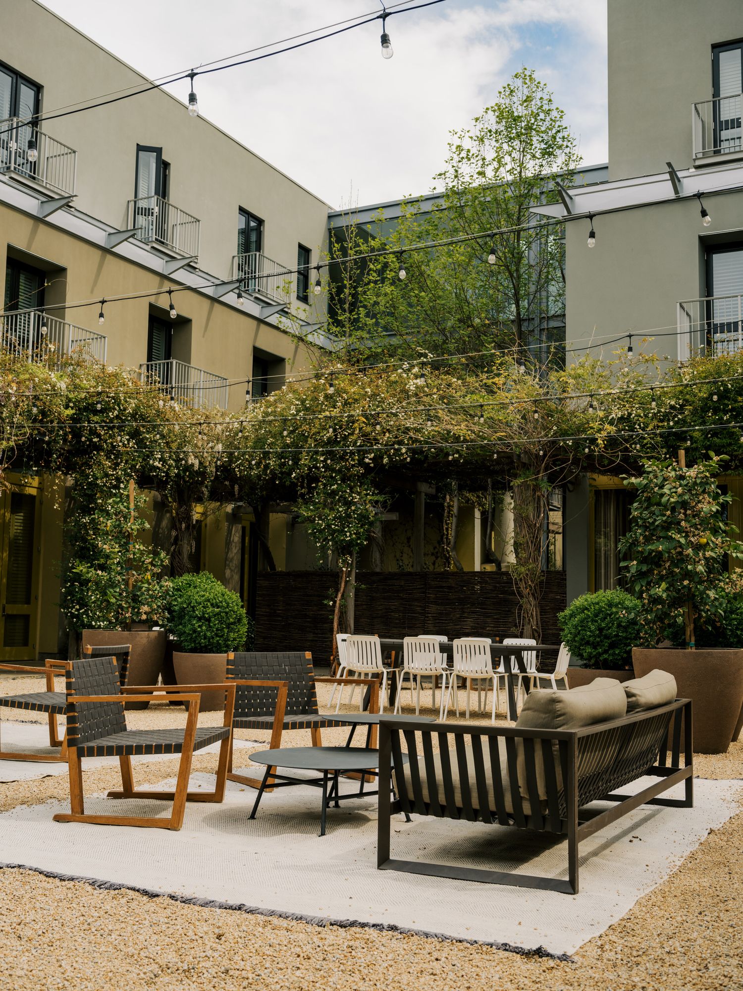 Modern outdoor patio space with multiple seating areas featuring wooden benches and chairs on gravel under string lights.