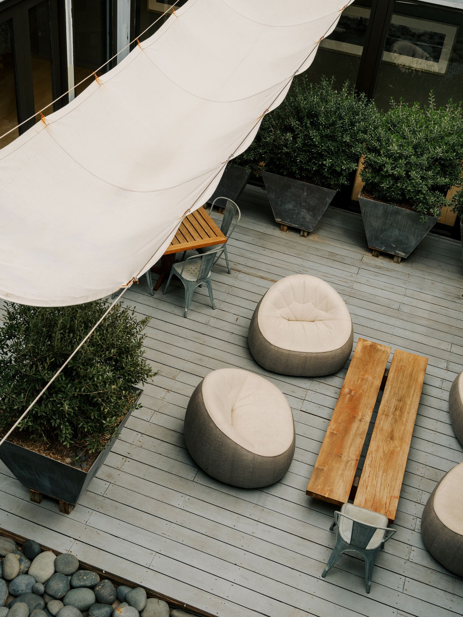 Modern outdoor deck space with gray planks featuring beanbag-style chairs, wooden bench and white sunshade canopy above.