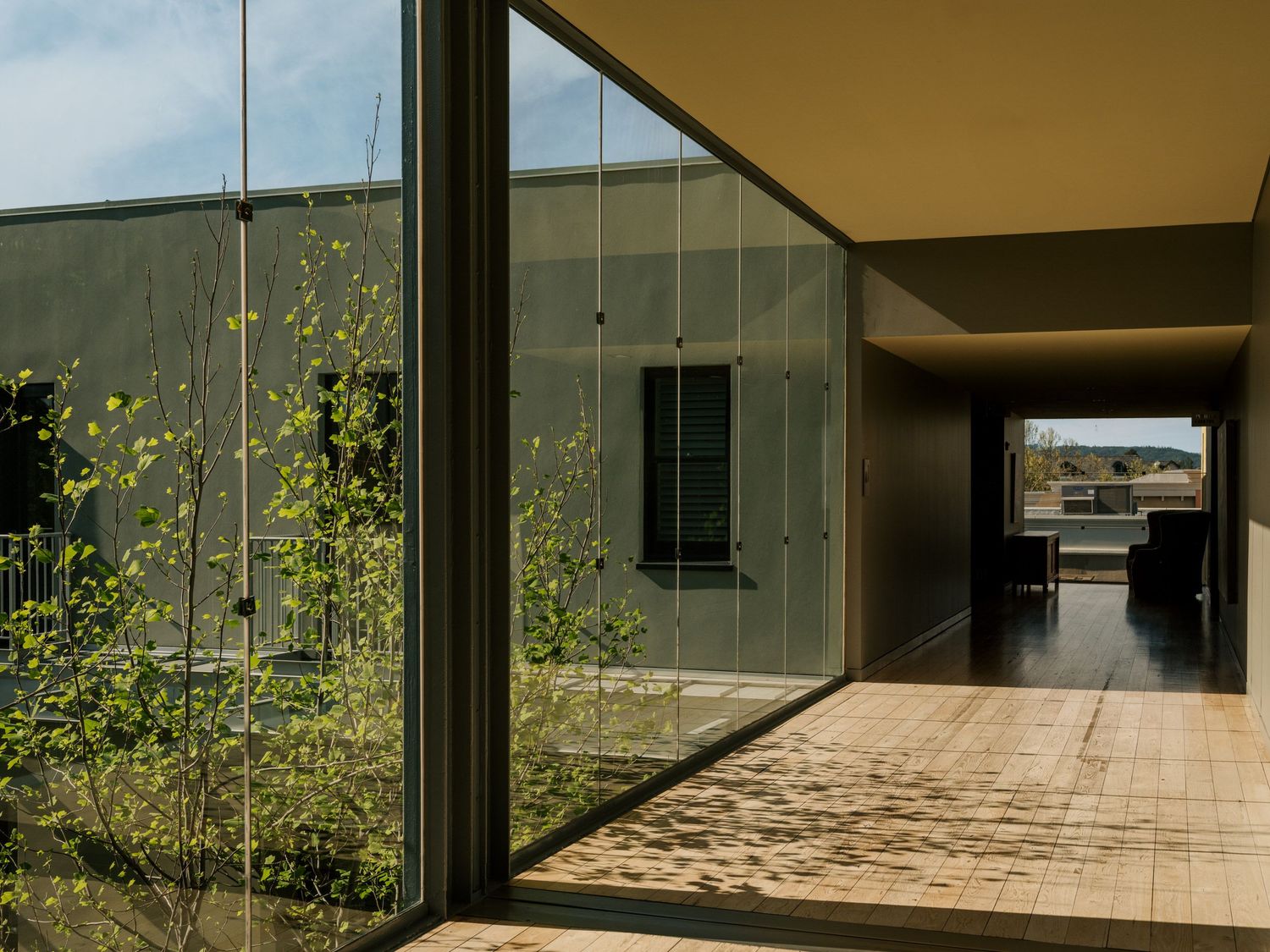Modern architectural walkway with floor-to-ceiling windows, glass walls and natural light reflecting on tiled flooring.