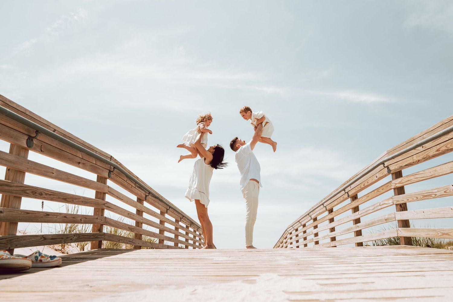 Family of four on a sunny beach boardwalk in Long Beach. Parents lifting two toddlers in the air with joy.