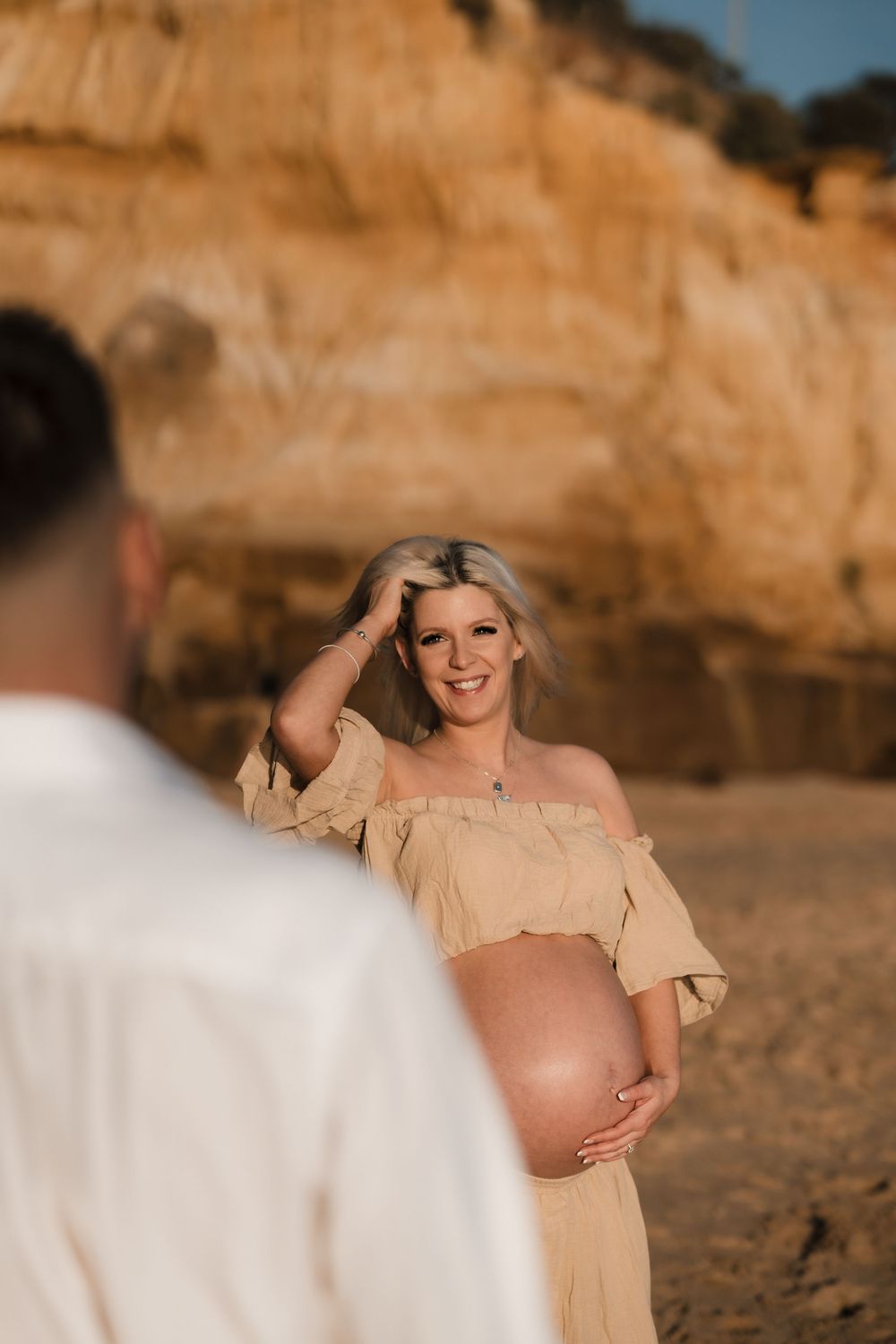 A maternity photoshoot at sunset features a flowing beige off-shoulder dress against dramatic coastal cliffs.