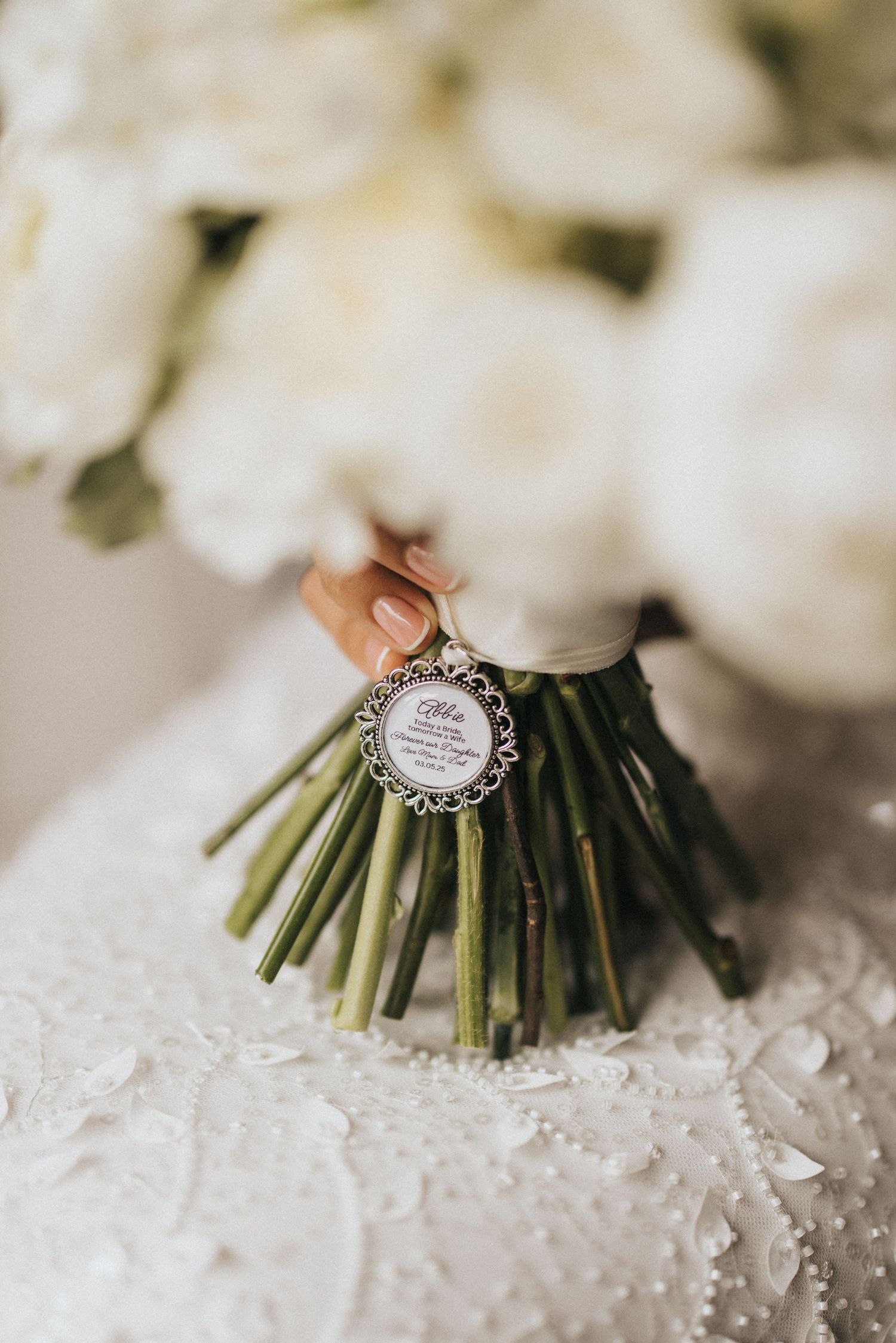 A wedding bouquet stem wrapped with a charm pendant and lace against a white background with soft bokeh.