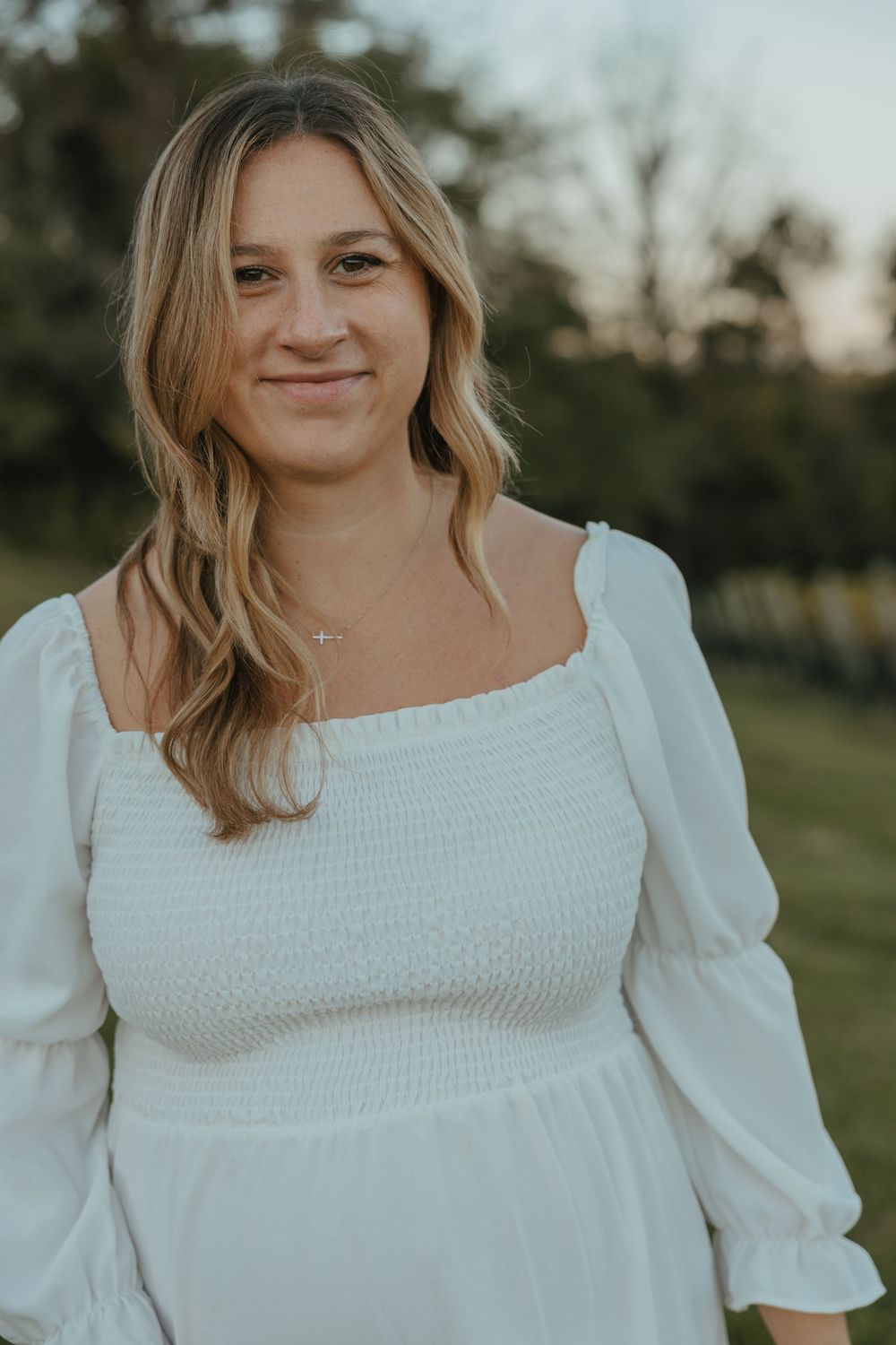 Woman in white smocked dress with puff sleeves stands outdoors during golden hour.