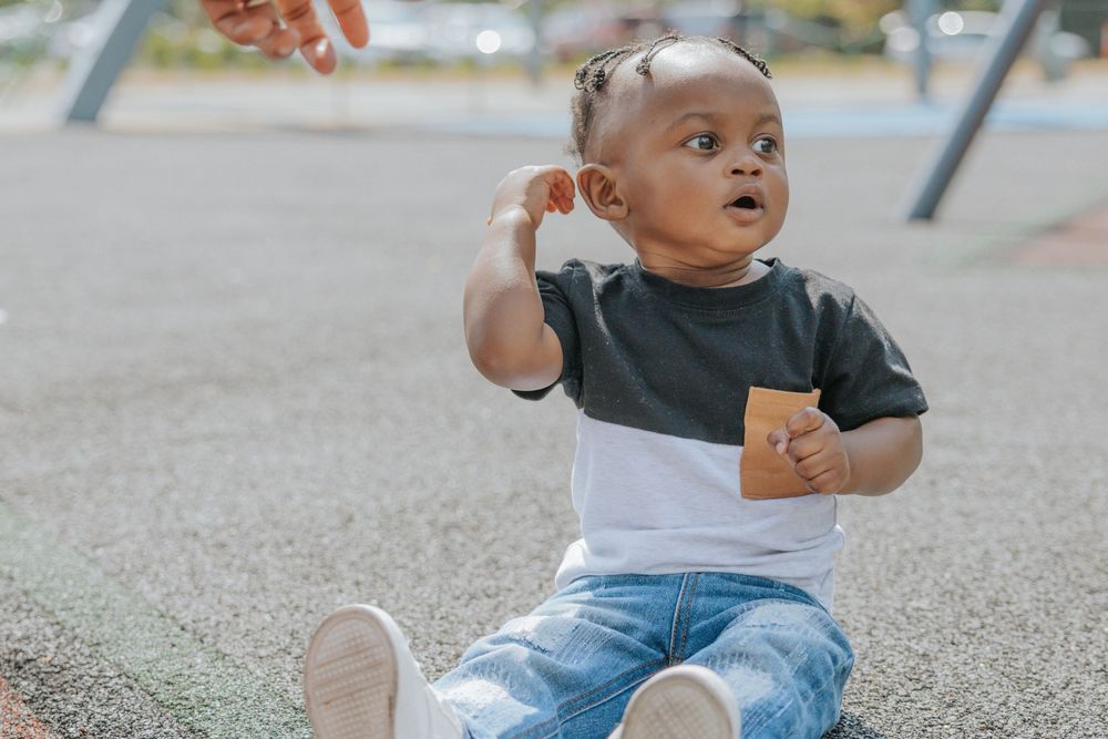 A baby in jeans and color-block shirt sits on a playground surface while looking up with curiosity.