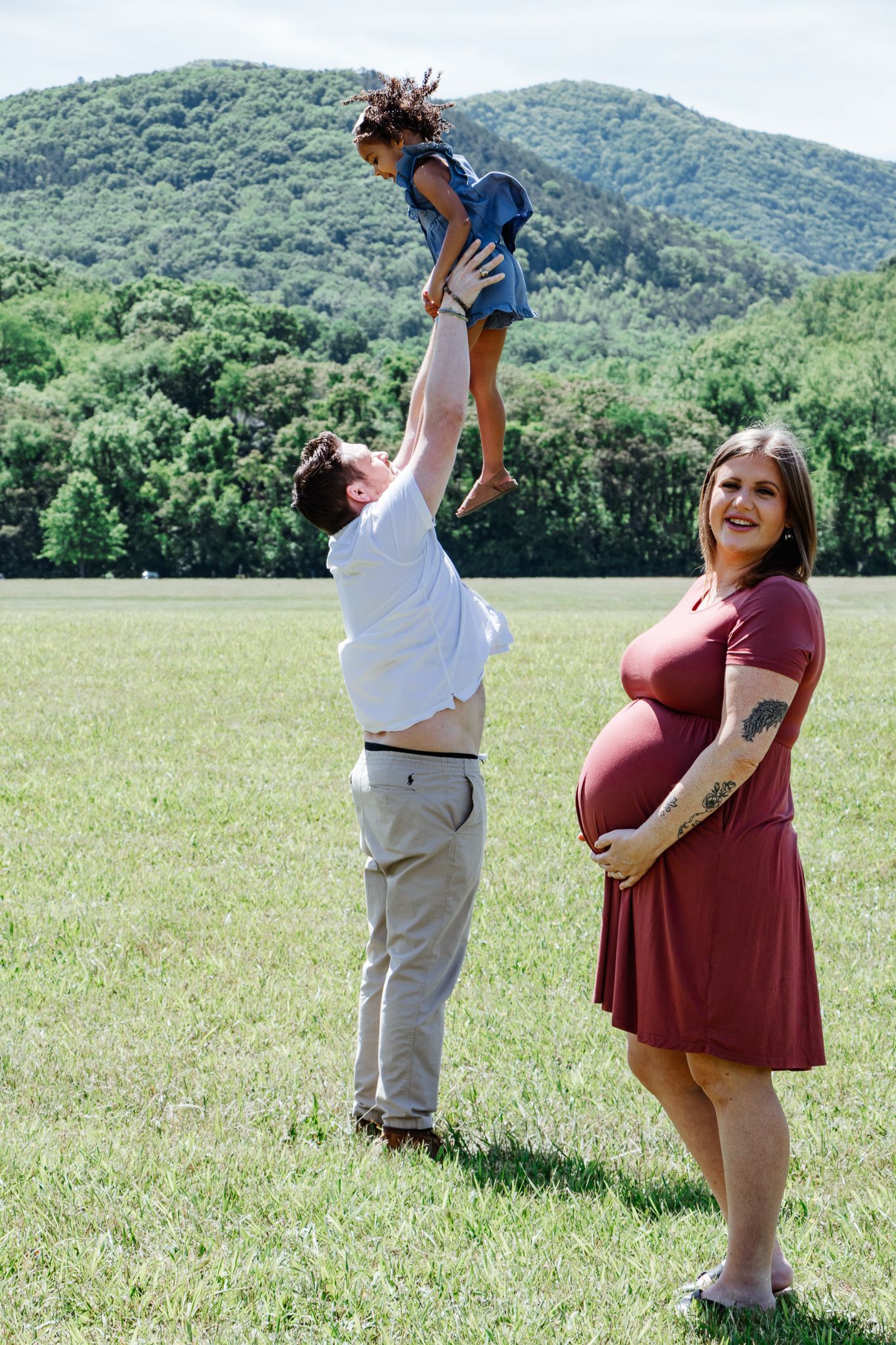Pregnant woman smiles while dad lifts daughter into the air in a sunny field with mountain and trees in the background.