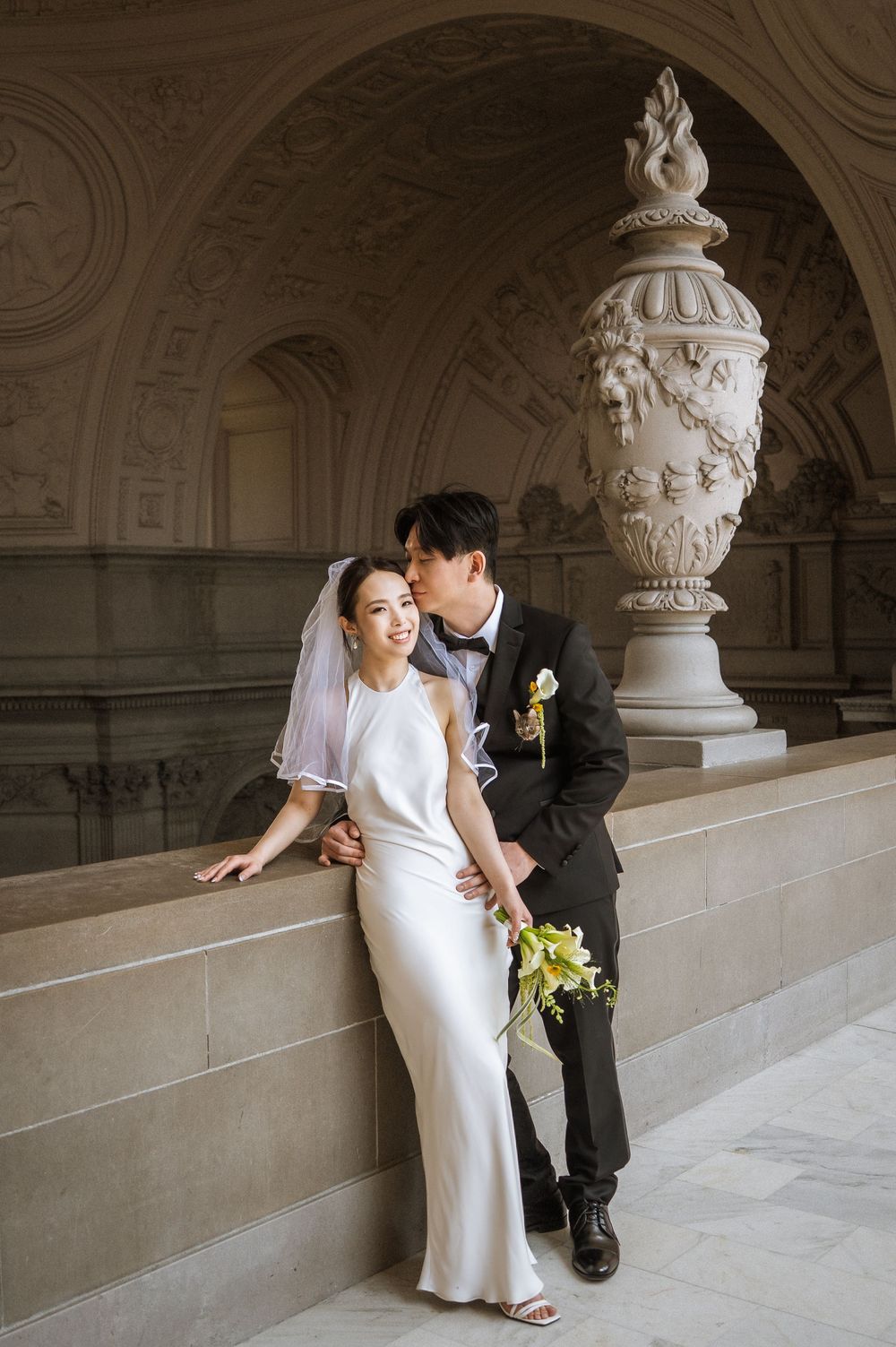Elegant wedding couple shares romantic moments in a grand marble hallway with ornate architectural details and columns.