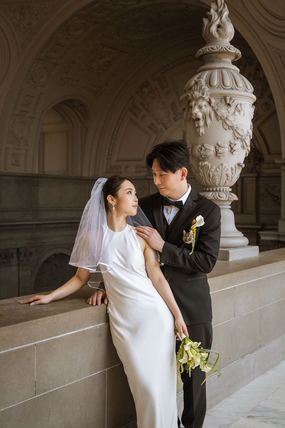 A couple in wedding attire pose romantically against an ornate stone archway with detailed classical architectural elements.