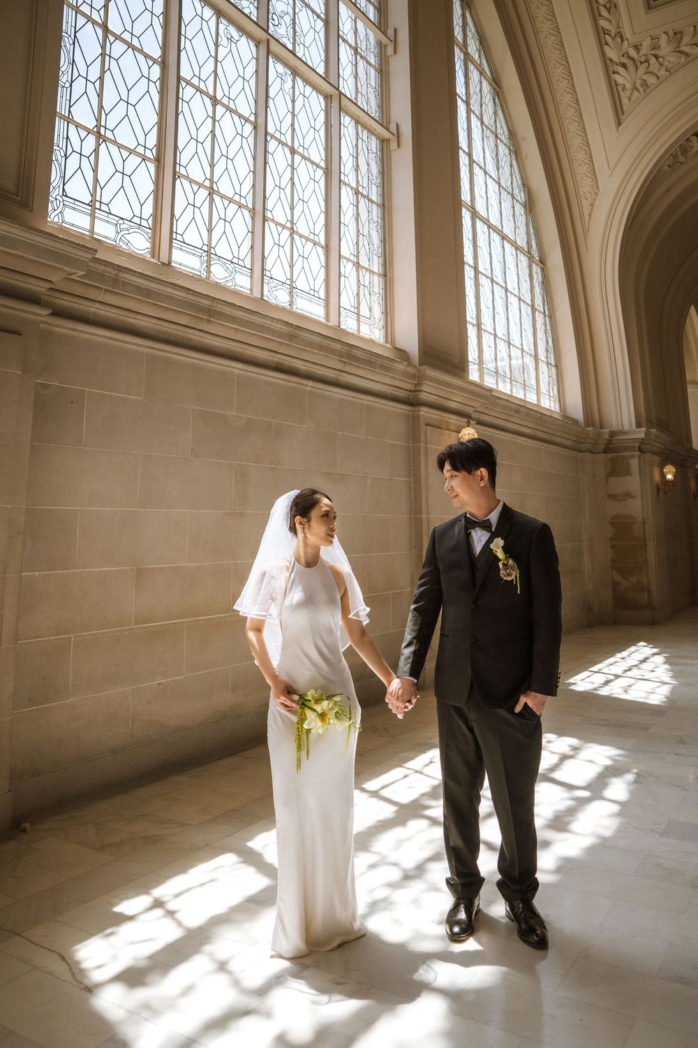 A couple stands together in a sunlit corridor with tall windows casting dramatic shadows on the stone floor.
