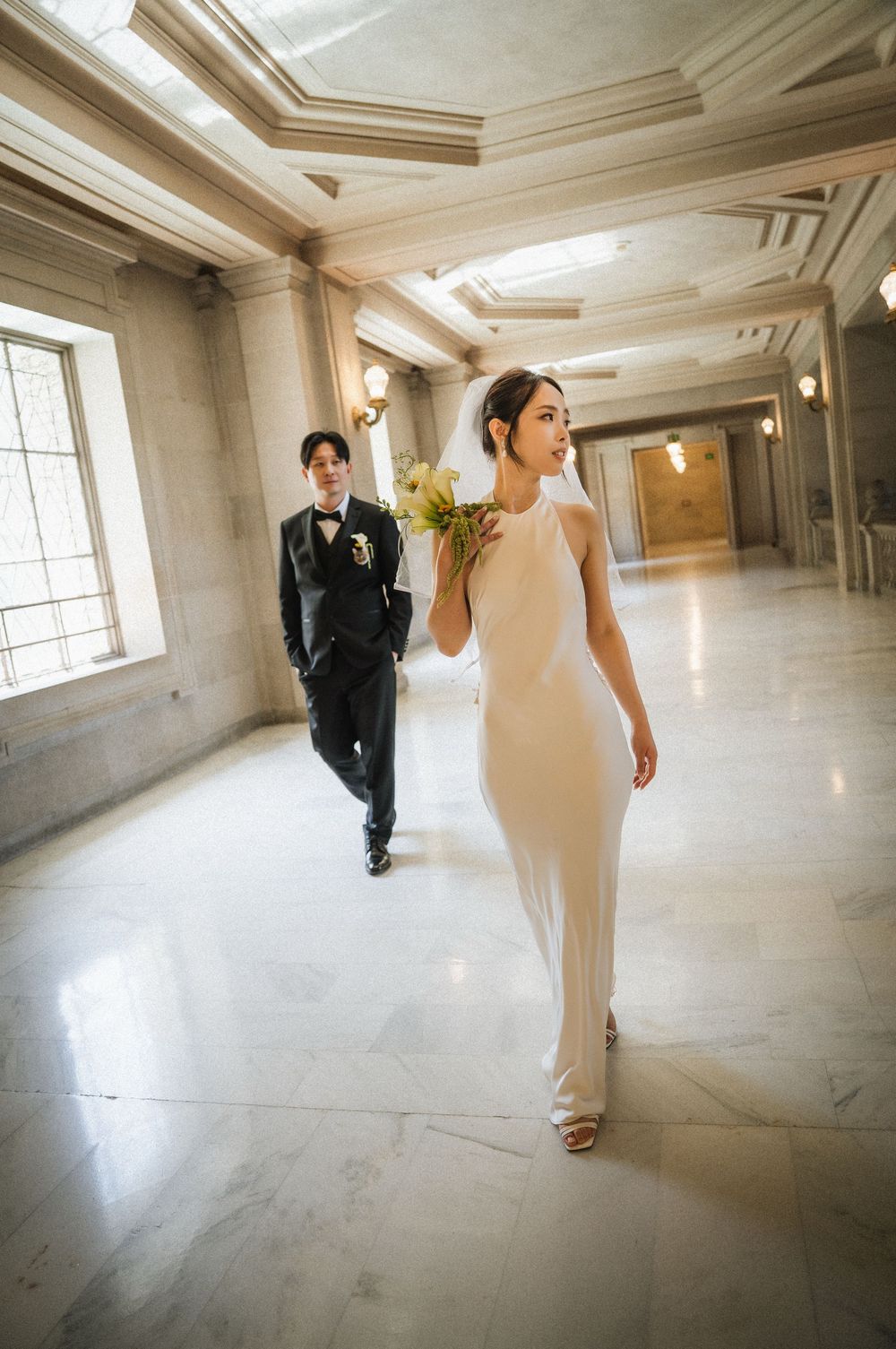 Elegant couple walks through a marble hallway with ornate architectural details at a formal wedding venue.