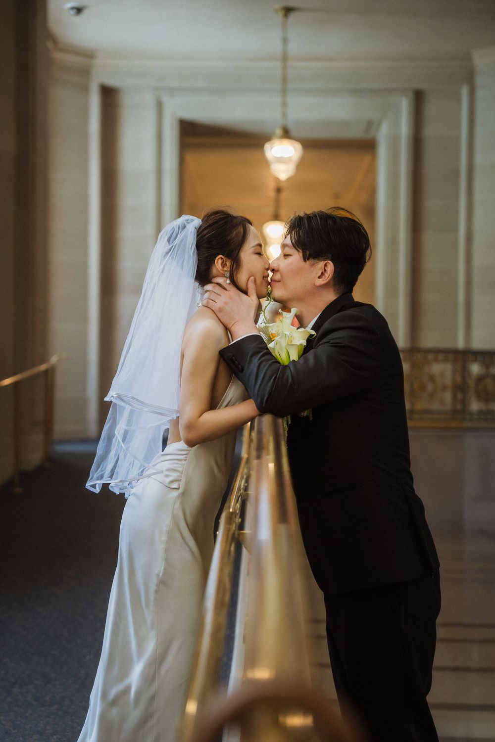A bride and groom share a romantic kiss in an elegant mansion hallway with warm lighting.