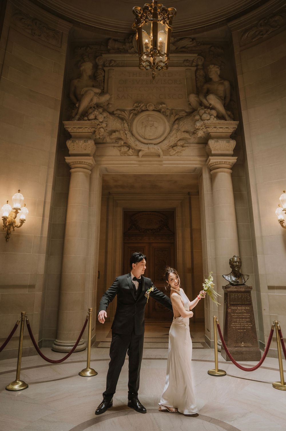A couple poses elegantly in a grand marble foyer with ornate architectural details, columns and antique light fixtures.